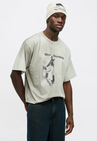 Φωτεινό πράσινο υπερμεγέθες βαμβακερό μπλουζάκι με γραφικό τύπωμα που αναγράφει "Won Hundred" και περιλαμβάνει μια εικόνα. Συνδυασμένο με σκούρο μπλε τζιν, φορεμένο με άνετο στυλ.