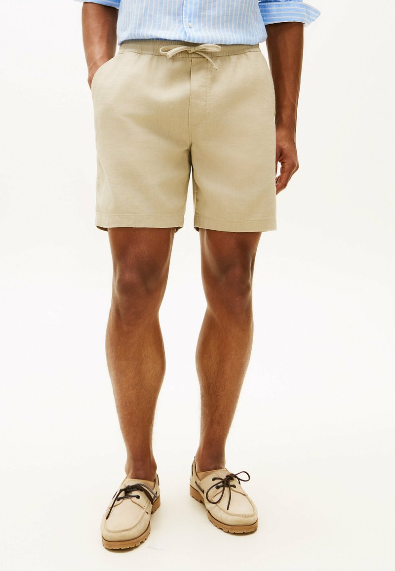 Homme portant un short beige à cordon, une chemise bleu clair aux manches retroussées et des chaussures bateau beiges à lacets, debout les mains dans les poches.