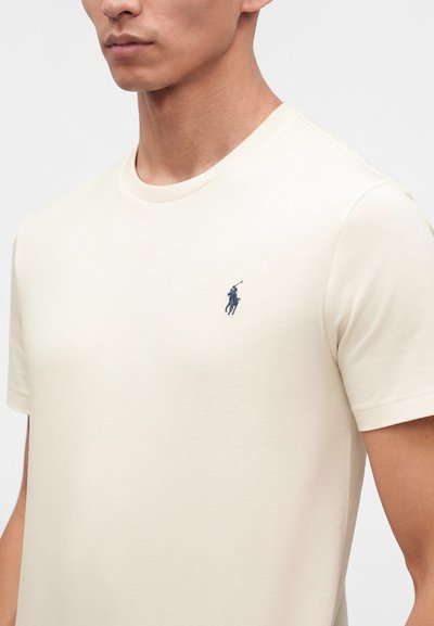 Άνδρας που φοράει απλό, κοντομάνικο, εκρού T-shirt με μικρό σκούρο μπλε λογότυπο παίκτη πόλο στο αριστερό στήθος σε λευκό φόντο.