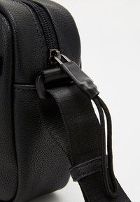 Borsa nera in pelle texturizzata con chiusura a zip, tracolla in nylon e tab in plastica. Presenta cuciture sottili e un design compatto.