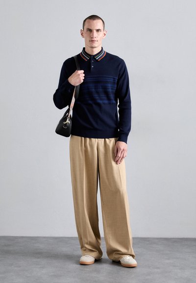 Navy knitted polo shirt with striped collar, paired with beige wide-legged trousers and light sneakers. Man holding a black crossbody bag.