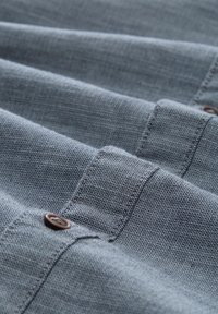 Plava tkanina u riblji uzorak s vidljivim šavovima i smeškim dugmadima, raspoređena u slojevite nabore, koja prikazuje detalje materijala i dizajna.