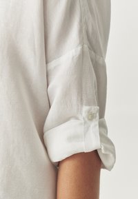 Camicia bianca di lino con maniche arrotolate. Presenta un dettaglio a bottone sul polsino. La trama sembra leggera e leggermente strutturata.