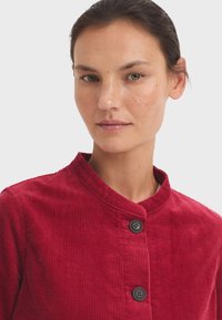 Rode corduroy jas met een ronde kraag en twee zwarte knopen. De stof heeft een ribbelstructuur en een aansluitend ontwerp.