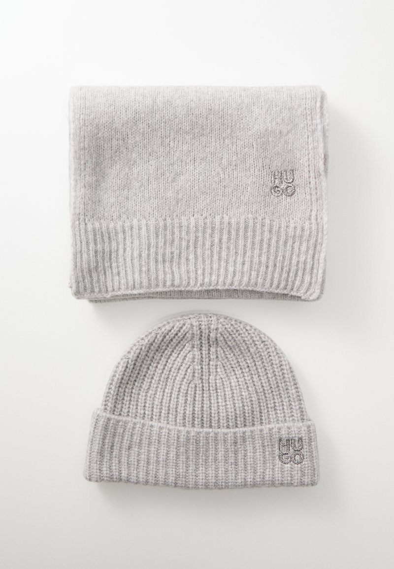 Gray knit scarf and beanie set, featuring ribbed texture and a small embroidered logo on the hat. Both pieces are made of soft fabric.