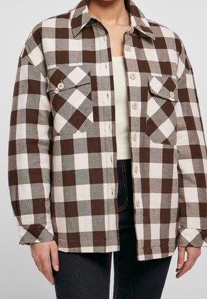 Femme portant une chemise à carreaux marron, blanche et grise avec deux poches à rabat sur le devant, par-dessus un haut beige et un pantalon foncé.