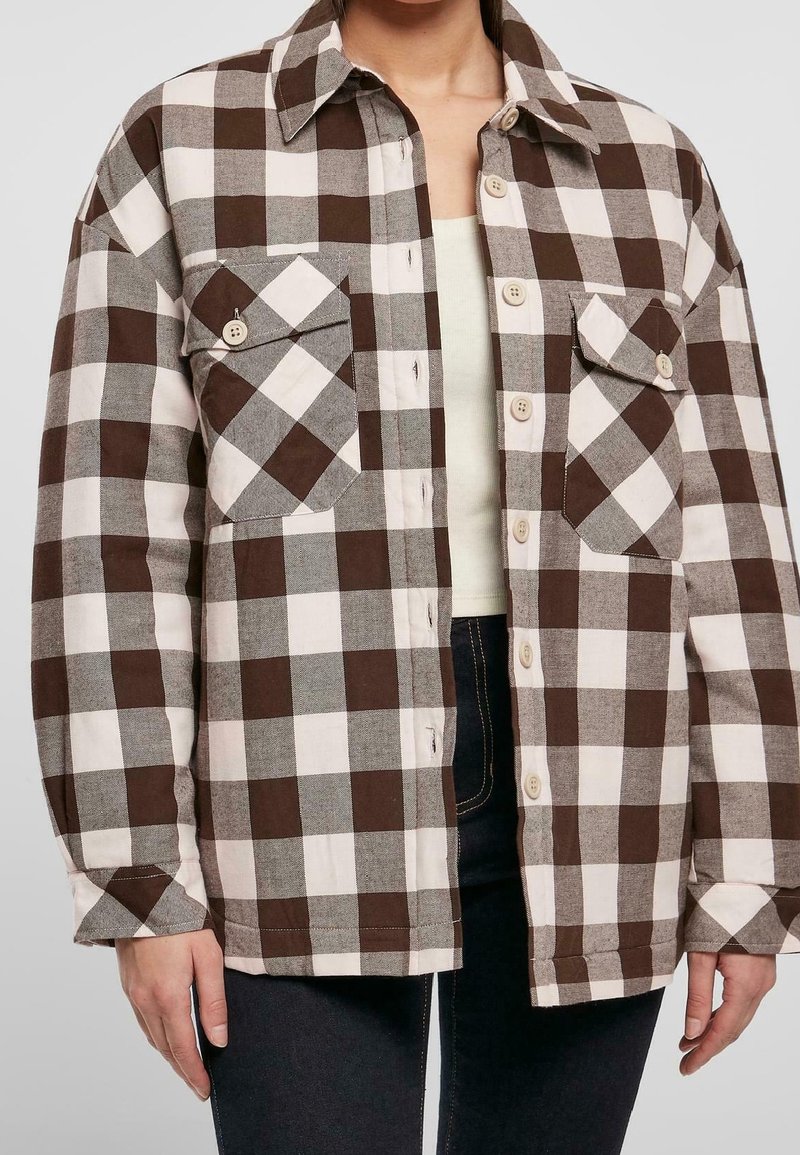 Femme portant une chemise à carreaux marron, blanche et grise avec deux poches à rabat sur le devant, par-dessus un haut beige et un pantalon foncé.