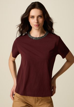 Donna con capelli lunghi e scuri che indossa una maglietta a maniche corte color bordeaux con una scollatura adornata di perline e pantaloni beige, in piedi davanti a uno sfondo semplice.