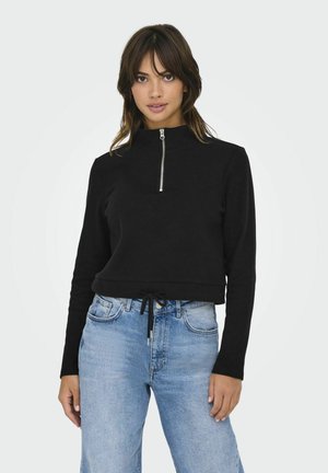 Sweatshirt noir court avec un col montant et une fermeture éclair demi-longueur, doté d'un ourlet à cordon de serrage. Assorti à un jean en denim bleu clair, légèrement déchiré.