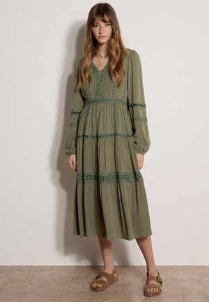 Femme aux longs cheveux bruns portant une robe midi olive à manches longues avec des détails en dentelle et des sandales marron à boucle, debout contre un mur blanc.