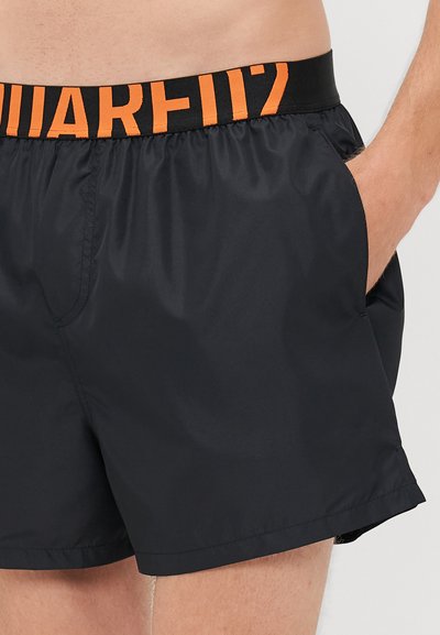 Schwarze Badeshorts mit elastischem Bund in Schwarz und orangefarbigem Text, getragen von einer Person mit den Händen in den Taschen.