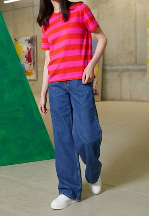 Person iført en pink og rødstribet t-shirt, brede blå jeans og hvide sneakers, der står indendørs nær et grønt trekantet kunstværk.