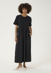 Juoda trumpų rankovių suknelė su laisvu kirpimu, turinti susiraukšlėjusį liemens detalę ir šonines kišenes. Lygus audinys ir kulkšnies ilgio.