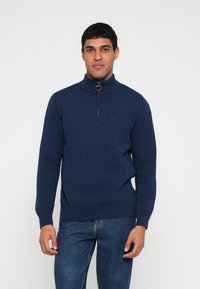 Camisola de malha azul-marinho com gola alta e fecho de correr de um quarto. Apresenta punhos e bainha canelados, com um detalhe sutil do logótipo.