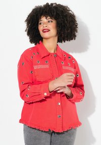 Chaqueta de mezclilla roja con dobladillo deshilachado, ojales plateados y tachuelas decorativas en los bolsillos. Dos bolsillos en el pecho y cierre de botones. Textura suave.