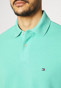 Pikétröja i mintgrön, tillverkad av texturerat tyg, med en krage med tre knappar och en liten broderad logotyp i röd, vit och blå.