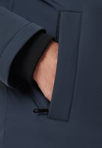 Ręka z czarnym mankietem w kieszeni granatowej kurtki, widoczny suwak.