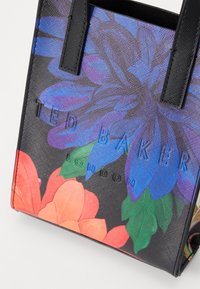 Črna torba s cvetličnim vzorcem, ki vsebuje živahne vijolične in oranžne cvetove. Vsebuje reliefni logotip "TED BAKER" ter močne črne ročaje.