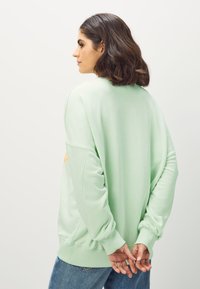 Mätovo zelená mikina s voľným strihom, nadmernými rukávmi, rebrovanými manžetami a zaokrúhleným výstrihom. Pohľad zozadu ukazuje jednoduchý dizajn bez ozdôb.