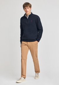 Navy fleece pullover met een halve rits kraag, voorzien van een klein rood logo, gecombineerd met beige chino's en witte sneakers. Zachte textuur.