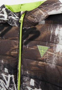 Giacca imbottita marrone con motivo astratto e strutturato. Presenta una cerniera verde e una fodera verde, con un logo triangolare sul petto.