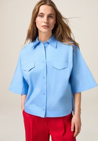 Camicia azzurra chiara a maniche corte con colletto, dotata di due tasche sul petto e orlo dritto, abbinata a pantaloni rossi. Texture liscia.