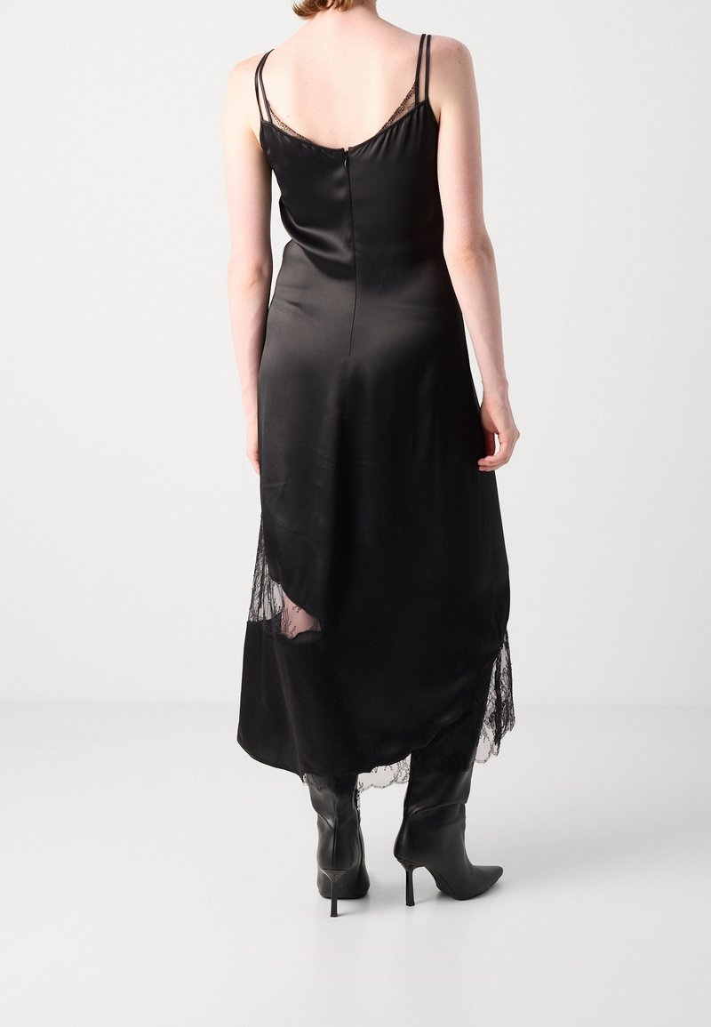 Femme portant une robe nuisette noire en satin avec bordure en dentelle et des bottes noires à talons hauts, debout de dos sur un fond uni.