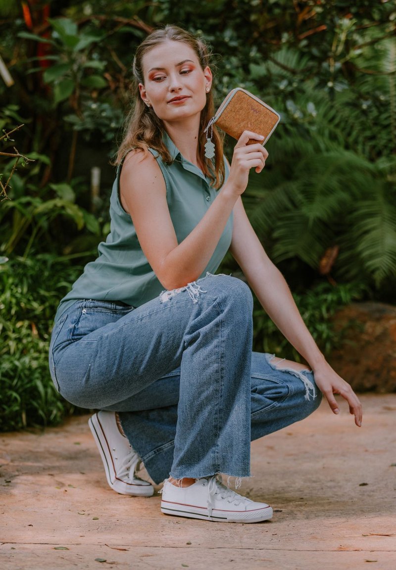 Cork wallet with a zipper, light brown texture, held by a person wearing a green polo shirt and distressed blue jeans with white sneakers.