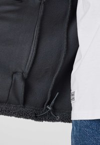 Veste en polaire noire avec ourlet réglable à cordon, portée sur une chemise blanche et un jean bleu, montrant l'intérieur de la veste et les détails de la texture.
