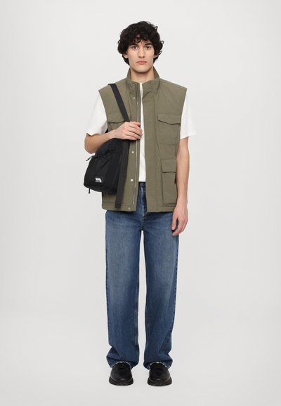 Gilet vert olive avec plusieurs poches, porté sur une chemise blanche et associé à un jean bleu à jambes larges et des chaussures noires. Sac bandoulière noir inclus.