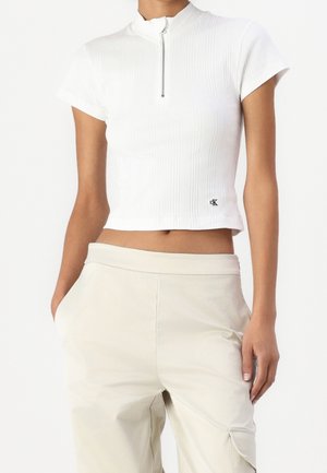 Mujer con una camiseta corta de manga corta acanalada blanca con cremallera frontal y pantalones beige de talle alto con bolsillos.