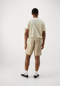 Camiseta de algodón beige y pantalones cortos beige a juego, con dos bolsillos traseros, combinados con zapatos negros y calcetines blancos hasta el tobillo.