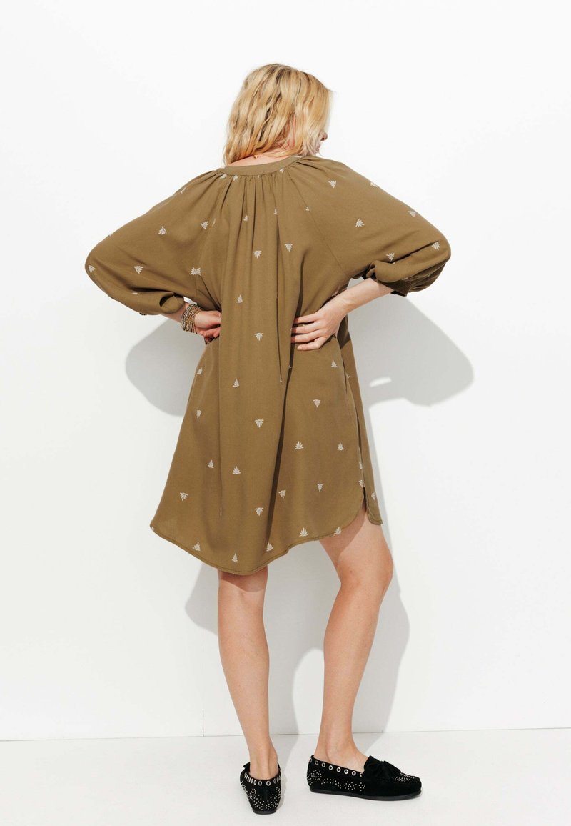 Robe vert olive avec des manches longues amples, arborant de petits motifs triangulaires blancs. Tissu doux tombant au-dessus du genou, ourlet arrondi, portée avec des chaussures noires.