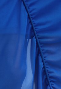 Šviesiai mėlynas tekstilas, kuriame derinamos vientisa ir permatoma medžiaga, su surinktomis raukšlėmis palei kraštą, kuriomis sukuriama tekstūra ir vizualinis susidomėjimas.