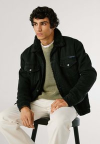 Chaqueta de mezclilla negra con un cuello de piel sintética, llevada sobre un suéter verde texturizado, combinada con pantalones color crema. Diseño suave y casual.
