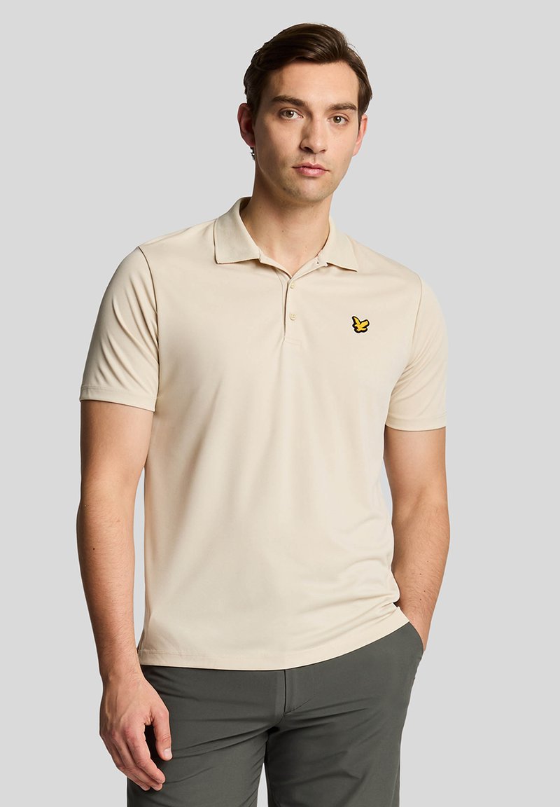 Beige polo skjorte med korte ermer, med krage og en gul sommerfugl logo på venstre bryst. Glatt stoff og tettsittende design.