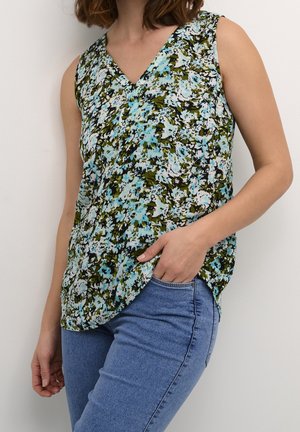 Frau trägt ein ärmelloses V-Ausschnitt-Oberteil mit floralem Muster in Blau, Grün und Weiß, kombiniert mit blauen Jeans, eine Hand in der Tasche, vor weißem Hintergrund.