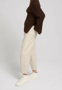 Osoba ubrana w kremowe spodnie z wysokim stanem, białe sneakersy za kostkę i ciemnobrązowy sweter z prążkowanego materiału z szerokimi rękawami, stojąca bokiem.