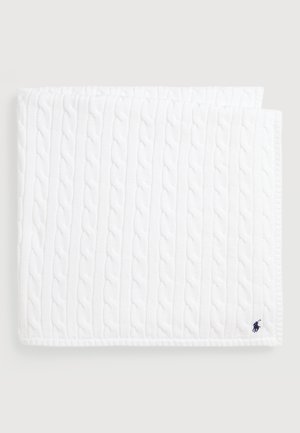 Valkoinen neulottu viltti, jossa on kaapelikuosi, teksturoitu pinta ja reunat. Laivastonsininen logo korostavana elementtinä alakulmassa.