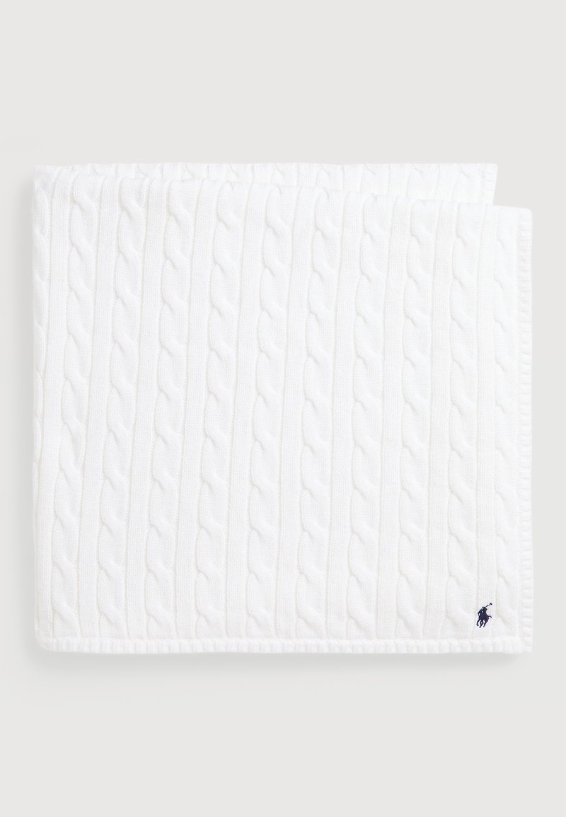 Baltā adītā sega ar kabeli rakstu, teksturētu virsmu un apmales malām. Logotipa akcenti tumši zilā krāsā apakšējā stūrī.