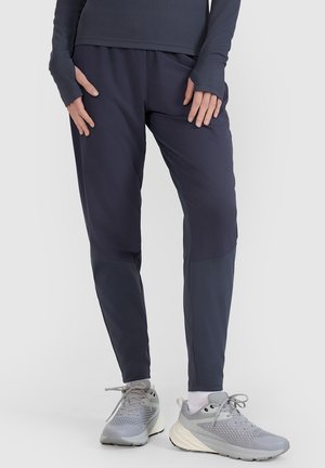 Personne portant un pantalon bleu marine foncé, des baskets grises et blanches, et un haut à manches longues foncé, debout devant un fond clair uni.