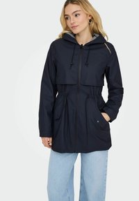 Veste imperméable bleu marine avec une capuche à cordon de serrage, des poches avant et une fermeture à boutons. Présente une coupe décontractée et une doublure intérieure contrastante.