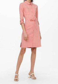 Abito rosa con colletto, due tasche sul petto e una cintura da annodare in vita. Le maniche arrotolate e le fessure laterali arricchiscono il design.