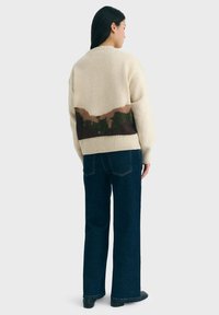 Woman wearing a cream sweater with brown and green abstract pattern on the back, dark blue jeans, and black shoes, standing facing away.