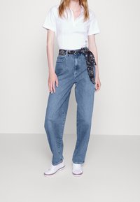 Vysoké modré džíny s volným střihem, doplněné černým páskem s potiskem, v kombinaci s bílými teniskami a bílým polo tričkem.