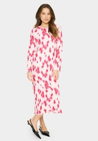 Rochie lunga, plisata, de culoare alba, cu modele vibrante de tie-dye rosu. Are maneci lungi si un decolteu rotund. Este asortata cu pantofi cu toc slingback negri.