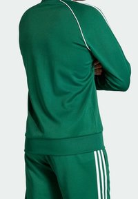 Chaqueta atlética verde con franjas blancas en los hombros y las mangas, hecha de un tejido suave y ligero, diseño ajustado.