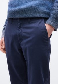 Άνδρας που φοράει ναυτικό μπλε παντελόνι και μπλε πουλόβερ με διαβάθμιση, με το ένα χέρι στην τσέπη, μπροστά σε μονόχρωμο φόντο.