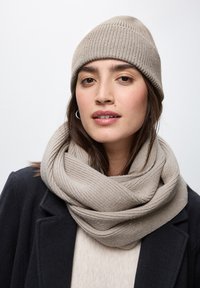 Cappello a coste beige e sciarpa oversize con un leggero luccichio, abbinati a un maglione crema e un cappotto scuro. Materiali testurizzati e accoglienti.