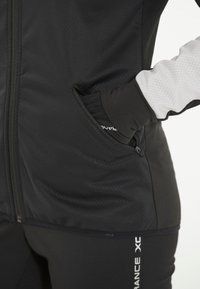 Schwarze Sportjacke mit einem Reißverschluss vorne und einer gemusterten Textur. Hat eine Seitentasche und kontrastierende graue Akzente an den Ärmeln.